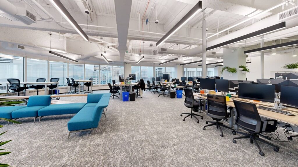 An open office space with modern furniture, including a blue couch and several workstations equipped with computers, surrounded by large windows.
