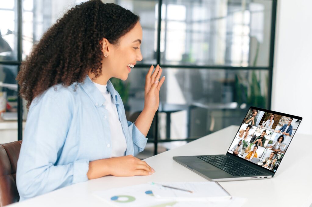 A woman with curly hair waves at her laptop while participating in a video call, showing a grid of people on the screen. She is seated at a desk with some papers and office supplies around her.