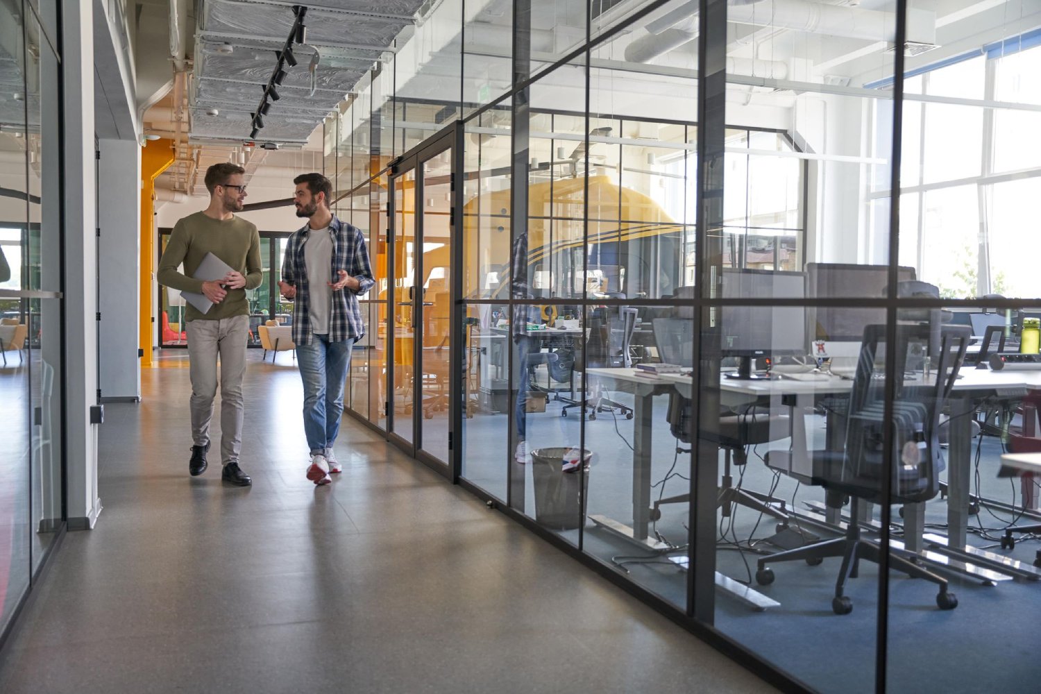 Two men walking and talking in a modern office space with glass partitions and empty workstations.