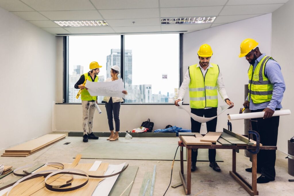 Four construction workers in hard hats and safety vests are inside an unfinished office space, reviewing plans and working with materials.