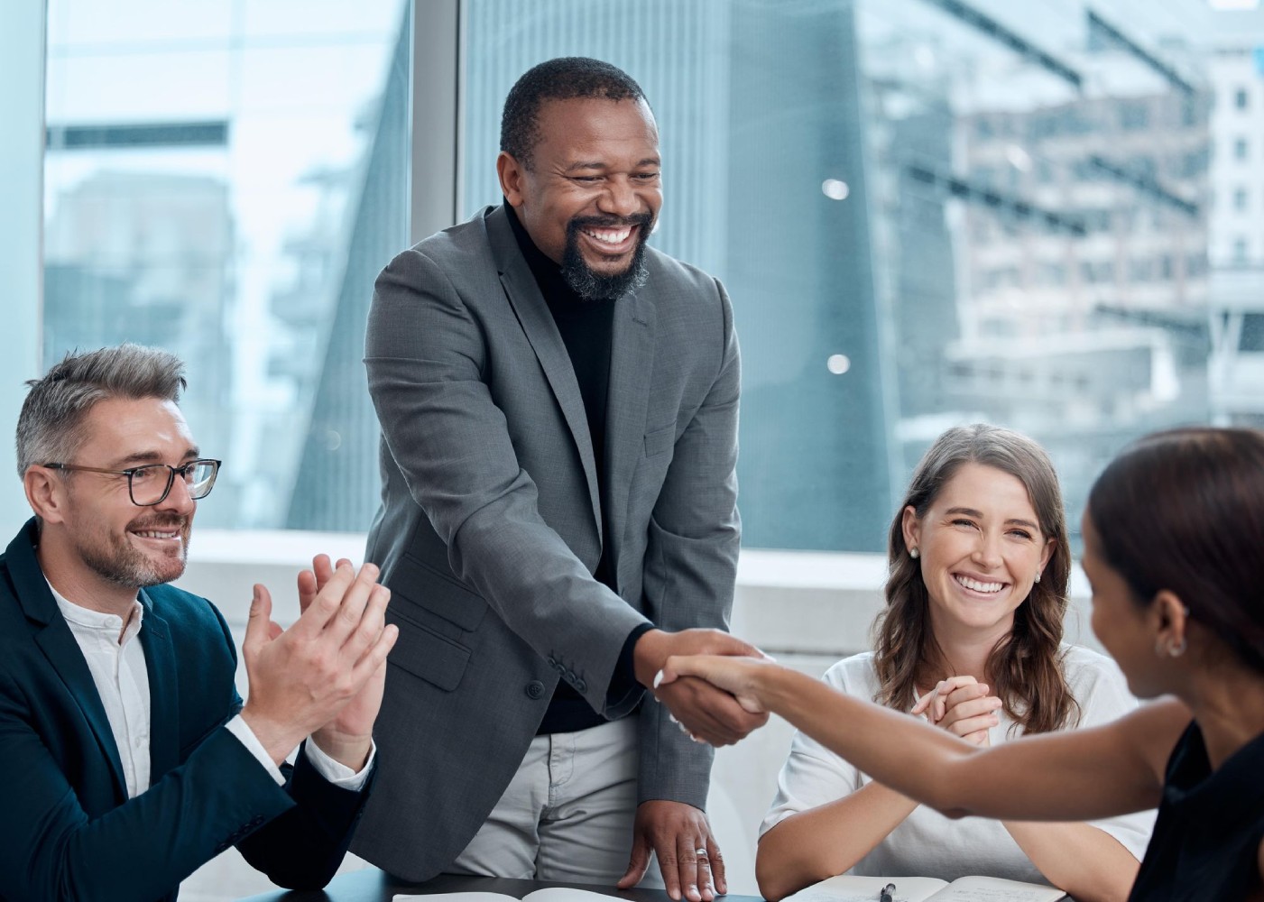 A group of professionals in a meeting, with a man shaking hands with a woman while others look on and smile.