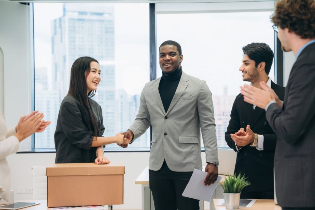A group of professionals in a modern office celebrating a successful handshake between a woman and a man, with others applauding in the background.