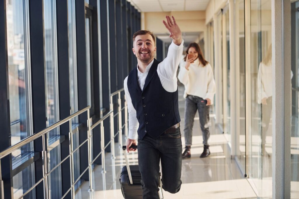 A man in a vest waves while walking down a corridor, pulling a suitcase, with a woman smiling in the background.