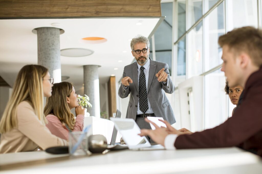 A businessman is standing and speaking to a group of four people sitting at a table in a modern office setting.