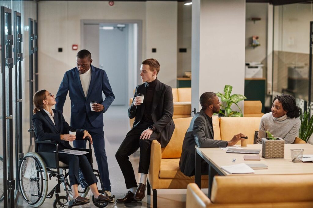 A diverse group of professionals engaging in conversation in a modern office setting. One person is in a wheelchair, while others are standing or sitting, holding cups.