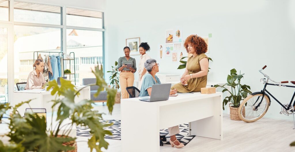A bright and modern workspace with several people engaged in various activities; one person is sitting at a desk with a laptop, another is seated nearby, and several others are standing or interacting in the background with plants and a bicycle present.