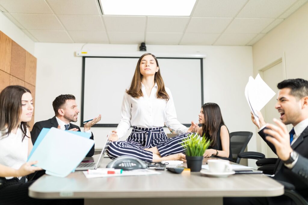 A woman sitting cross-legged on a conference table, meditating, while four colleagues around her appear to be arguing or stressed in a chaotic office meeting.