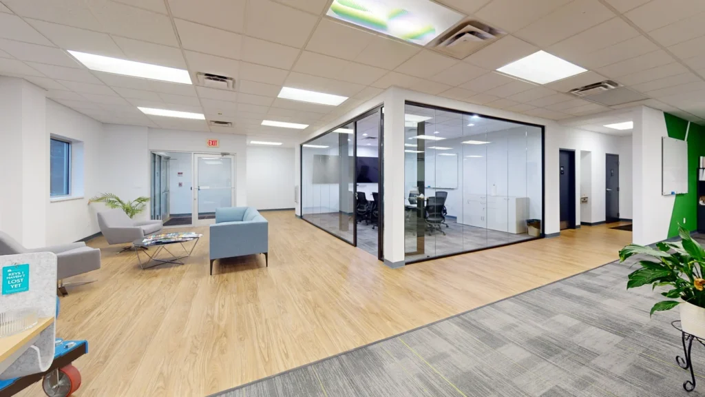 Open office lobby and reception area featuring wood-look flooring, glass-enclosed conference rooms, comfortable lounge seating with a blue sofa and grey armchairs, and a small coffee table.