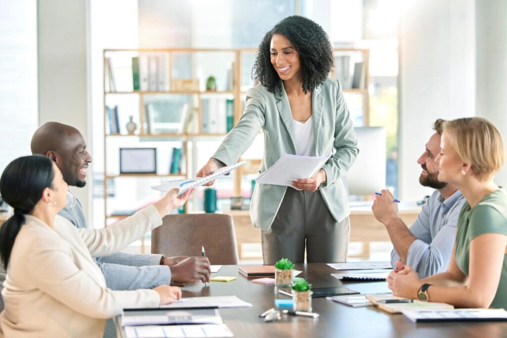 A woman stands at a podium, presenting to a group of attentive colleagues seated in a conference room.