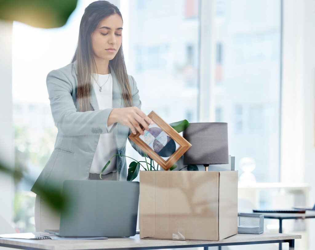 A woman in a professional outfit is packing a box with personal items, including a picture frame, at a desk with a laptop and plants.
