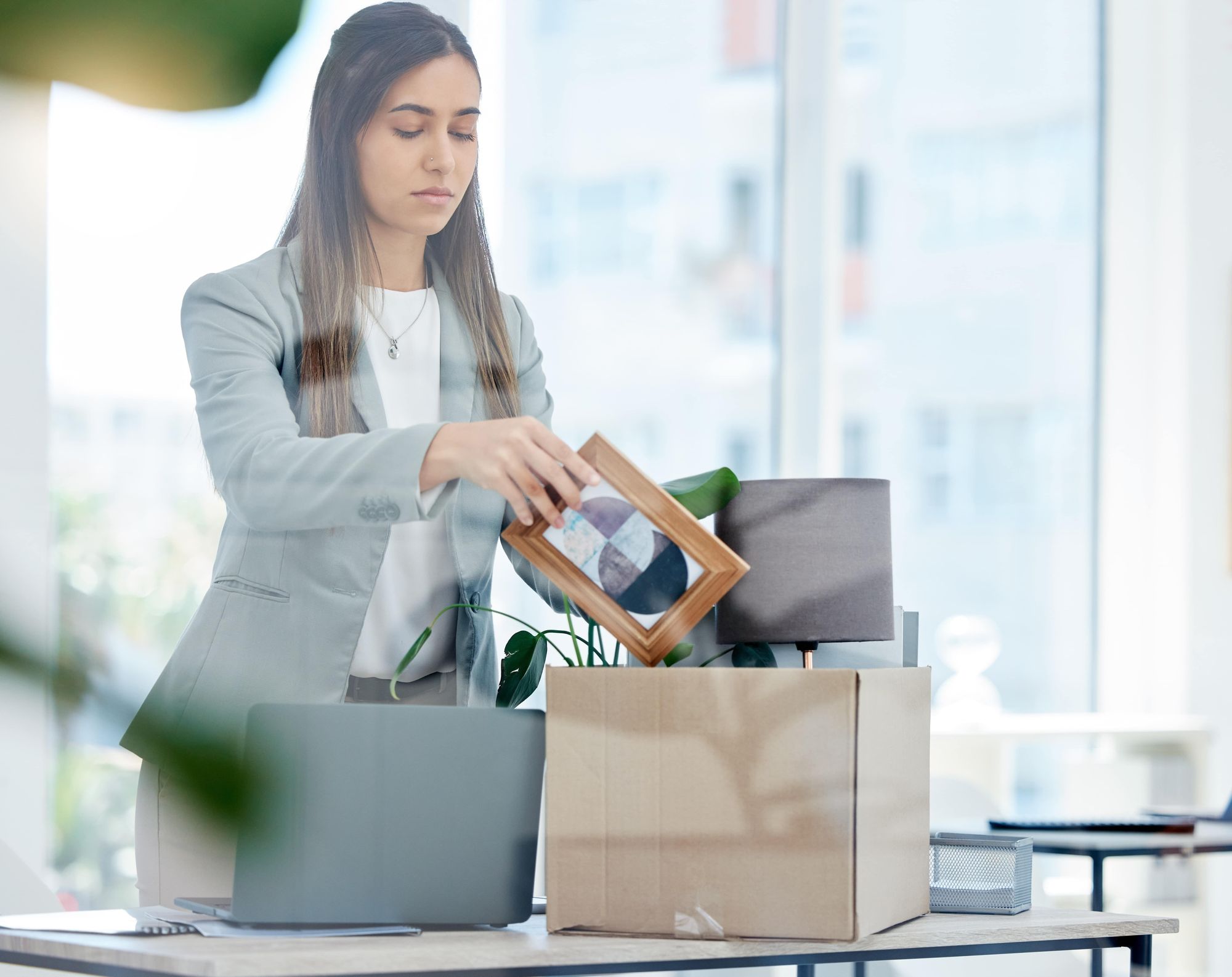 A woman in a professional outfit is packing a box with personal items, including a picture frame, at a desk with a laptop and plants.