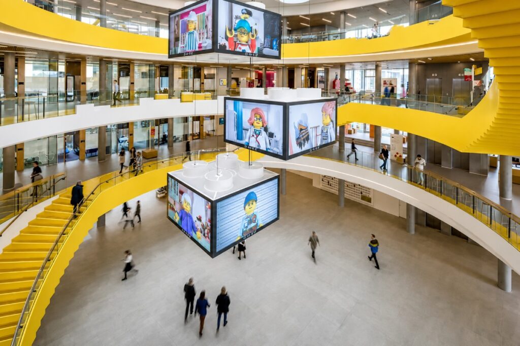 A modern atrium with yellow accents, featuring hanging digital displays and people walking around the open space.