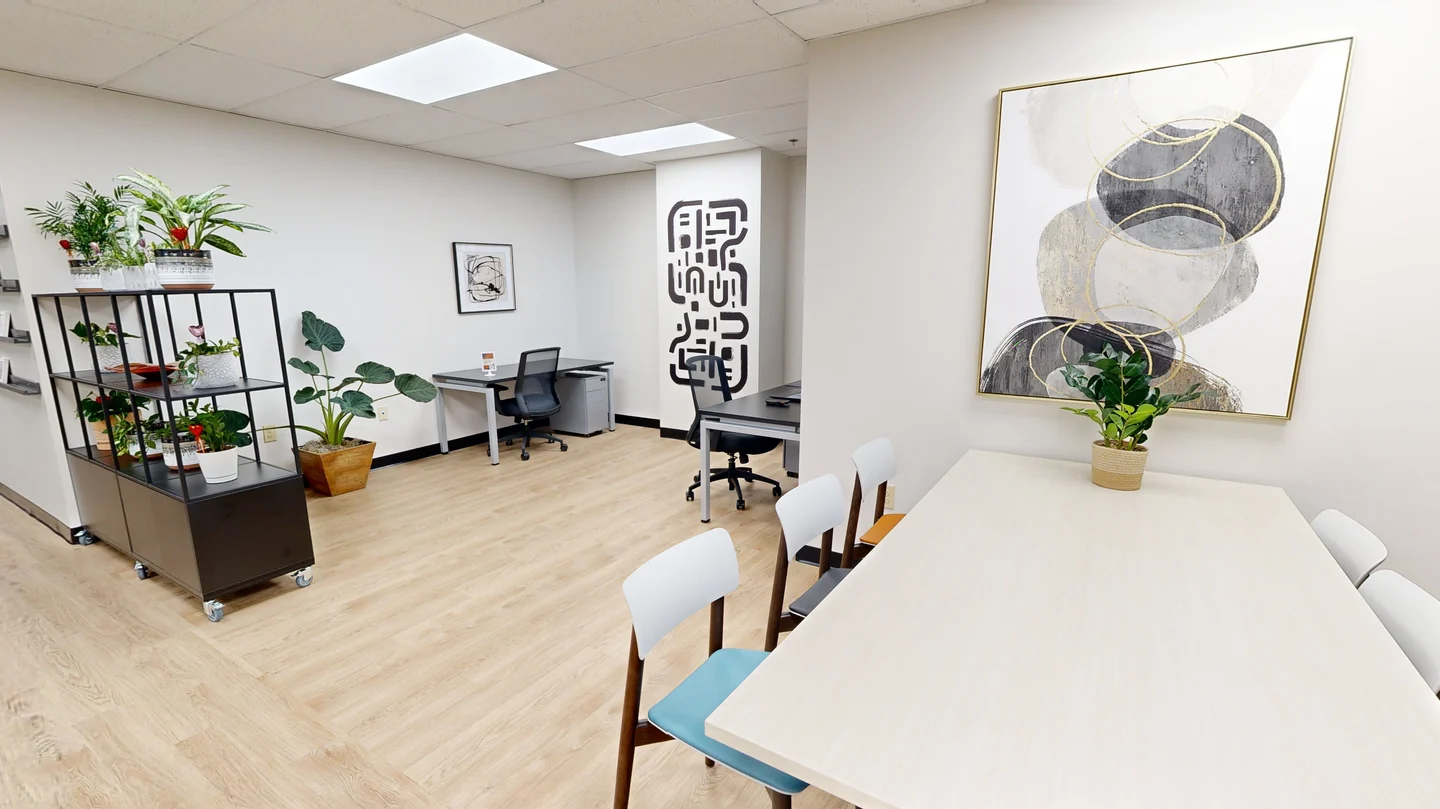 Bright, modern open-plan office lounge and co-working area with light wooden floors, potted plants, a multi-tier display shelf, individual desks, and a dining table with chairs.