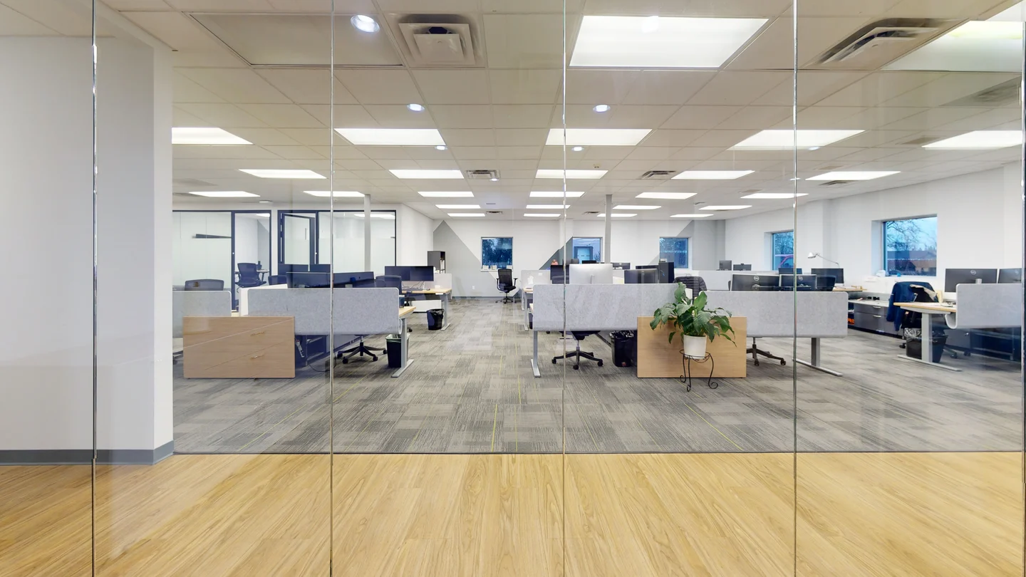 Wide-angle view of a large, bright open-plan office seen through floor-to-ceiling glass walls, featuring carpeted workstation rows with privacy partitions and office chairs.