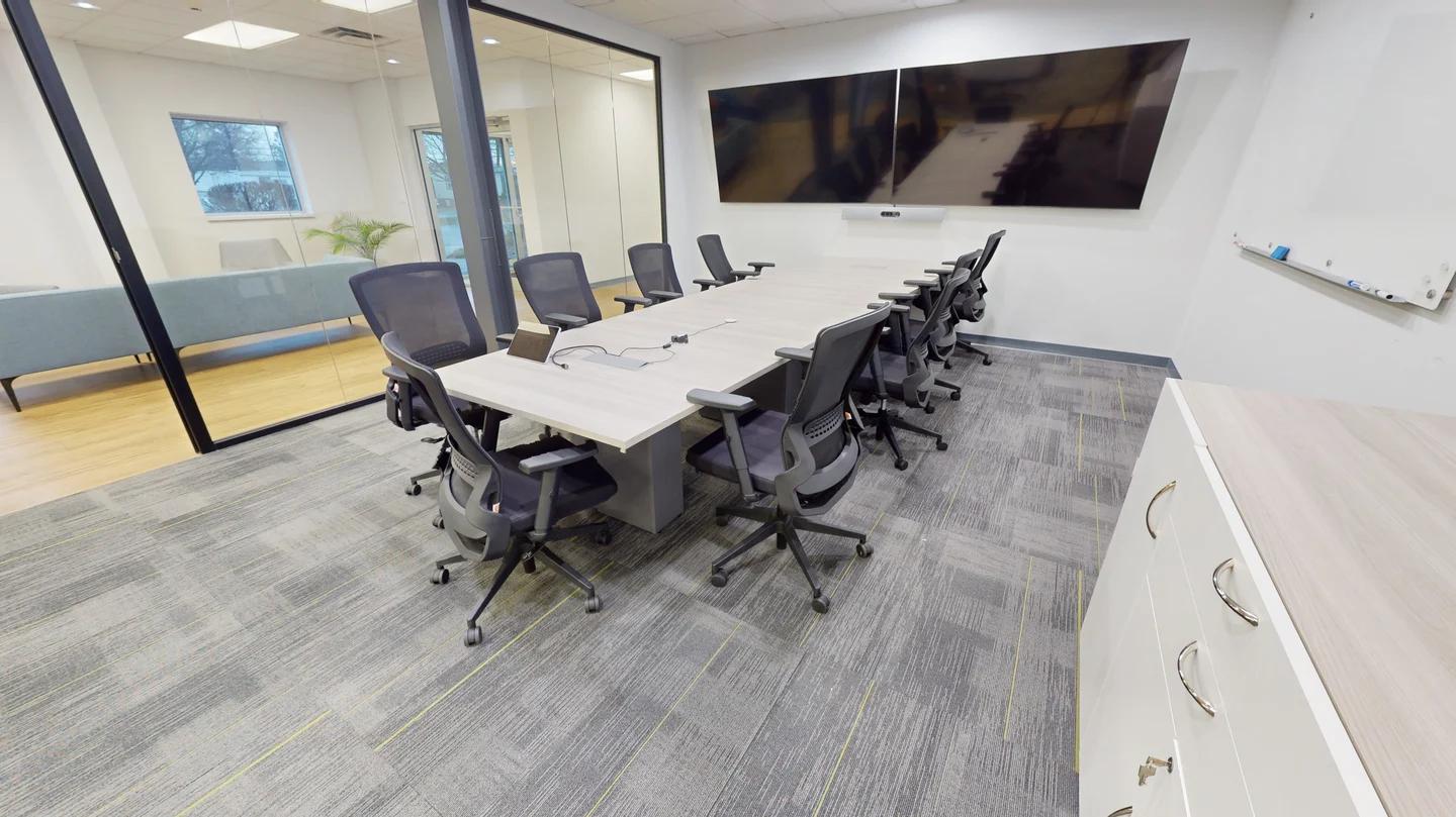 Modern conference room featuring a long light-wood table surrounded by black mesh office chairs, dual wall-mounted presentation monitors, carpeted flooring, and glass wall partitions.