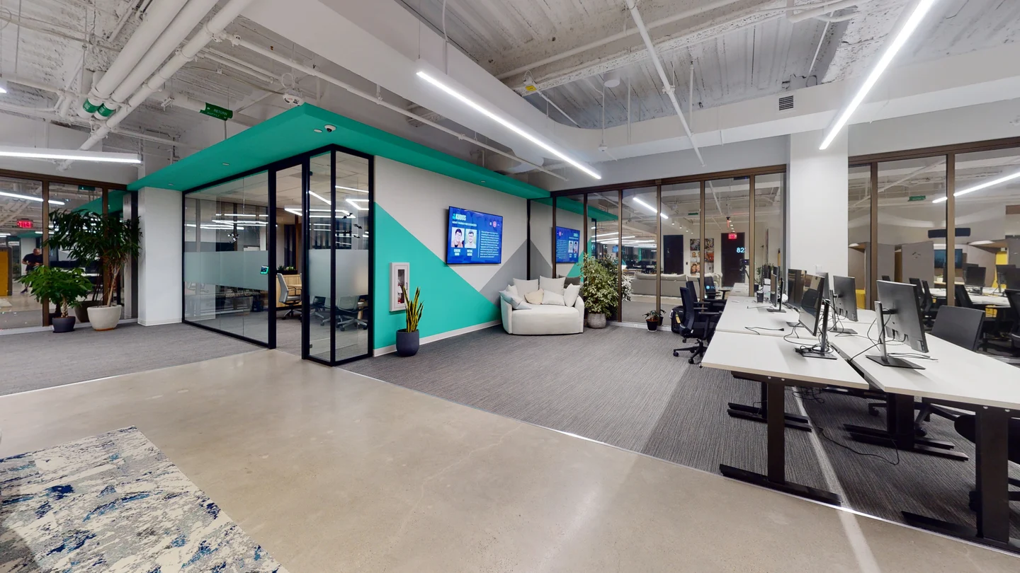 Contemporary open office layout featuring modern computer desks with dual monitors, a glass-enclosed meeting room, vibrant teal accent walls, and exposed industrial ceiling pipes.