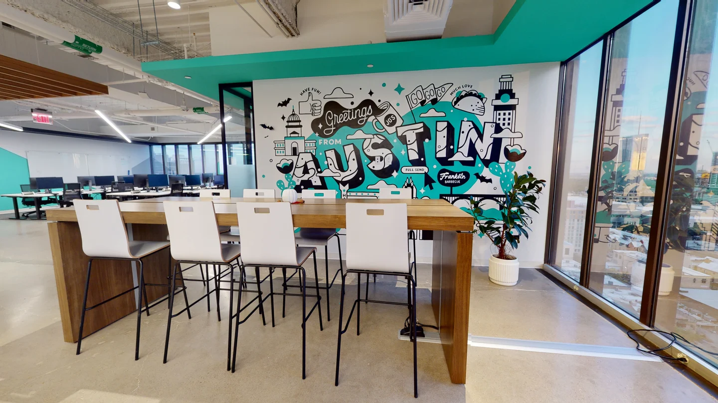 Modern office break area featuring a high-top wooden table with white bar stools set in front of a colorful "Greetings from Austin" wall mural and floor-to-ceiling windows.