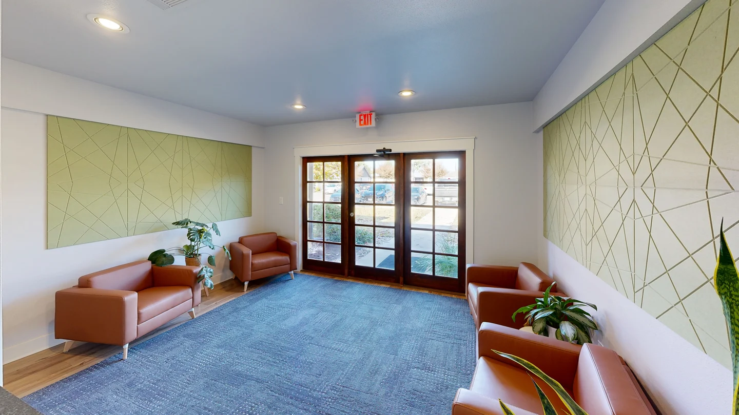 Office reception waiting area with leather armchairs, blue carpet, potted indoor plants, light green textured wall panels, and glass entrance doors.