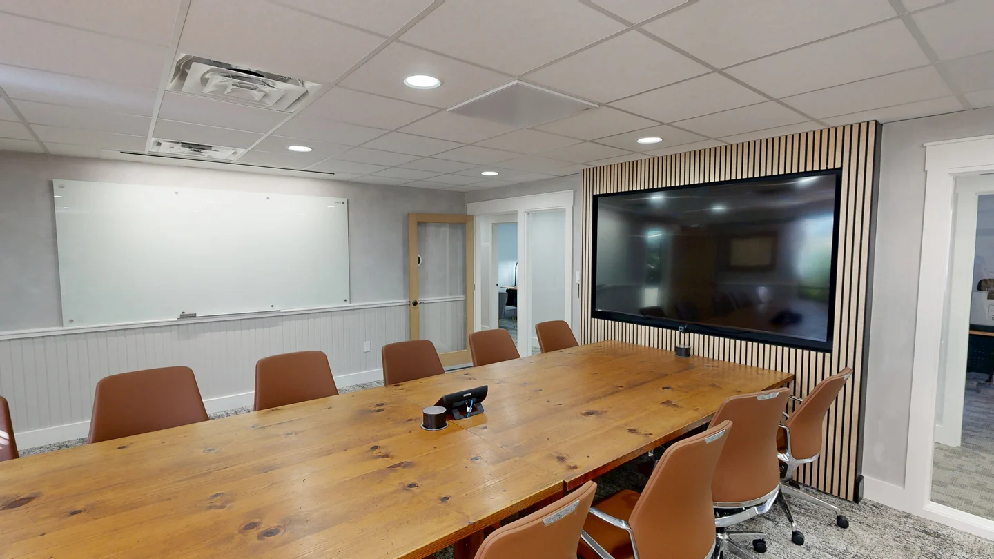 Modern board room with a large wooden conference table, brown leather executive chairs, a large display monitor mounted on a wood-slat accent wall, and a large whiteboard.